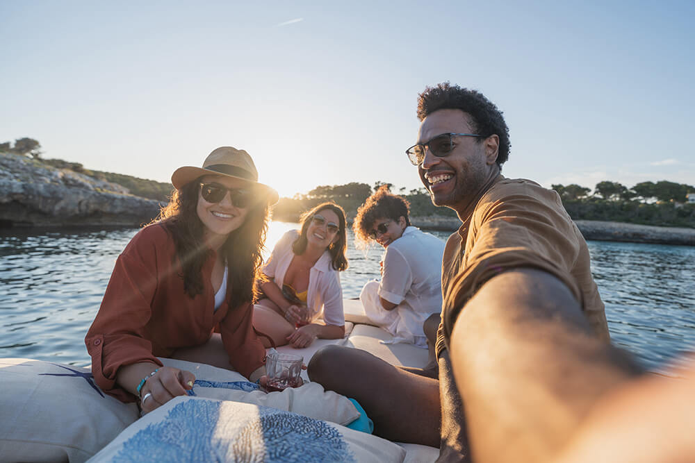 Four friends smile and pose for a selfie while sitting on a boat, enjoying a sunny day on the water with rocky shores and trees in the background.